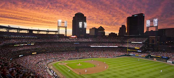 An orange and purple sunset lights the sky above Busch Stadium during a baseball game in St. Louis, Missouri