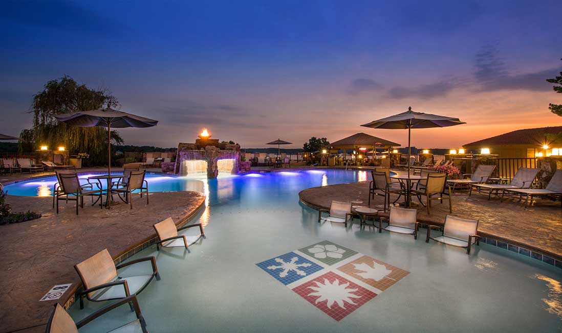 A lakeside pool at sunset, with in-pool lights and chairs, surrounded by round tables with chairs and umbrellas on the deck at Lodge of Four Seasons in Lake of the Ozarks, MO