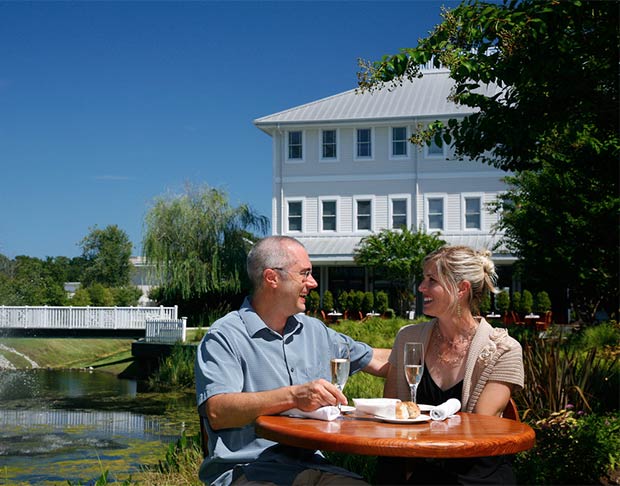 A couple sits at a round outdoor table with glasses of champagne, in front of a white building and overlooking a white bridge over water at Lumina Station in Wilmington, North Carolina