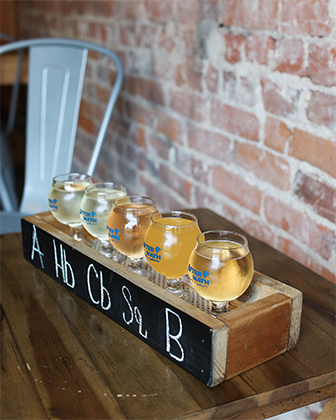 A wooden box with chalk labels on a table holds glasses of cider in front of a brick wall in Indiana Dunes, Indiana