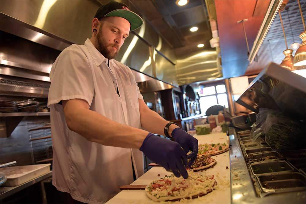 A pizza chef wearing a baseball cap adds toppings on to a pizza on the prep line at Chiusano’s Brick Oven Pizzeria in Kansas City, KS