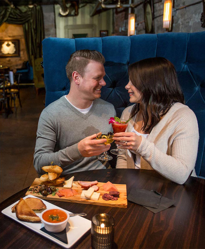 A man and woman holding drinks look into each other’s eyes while seated in a blue booth with a table that has a platter of meat and cheese as well as a sandwich on a plate and a bowl of soup at The Velveteen speakeasy in Stillwater, MN.