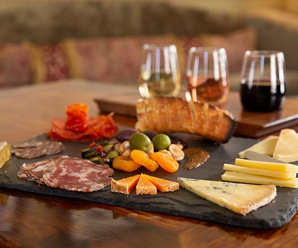 An extreme close up of a charcuterie tray with various cheeses, meats, bread and an olive and nut mixture. Behind the tray is a flight of wine with three glasses: white, rose and red. 