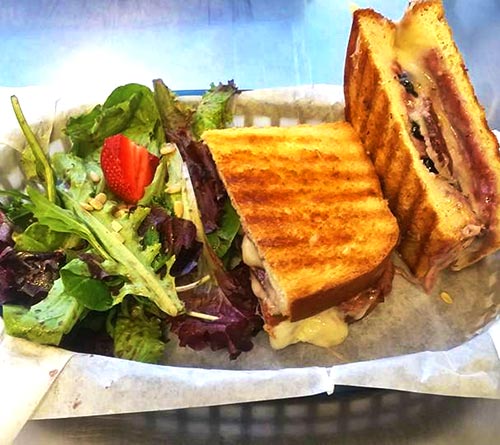 An extreme close up of a blueberry grilled cheese sandwich cut in half, made with applewood smoked bacon, havarti, berry aioli, and blueberries grilled on sourdough served with a side salad in a plastic basket.