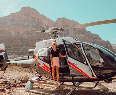 A woman wearing sunglasses stands in the open door of a helicopter on the ground at the Grand Canyon
