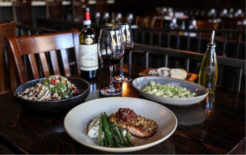 A plate of crusted salmon and asparagus sitting on a bed of mashed potatoes. Some appetizers, and another dish sits with a bottle of Italian red wine