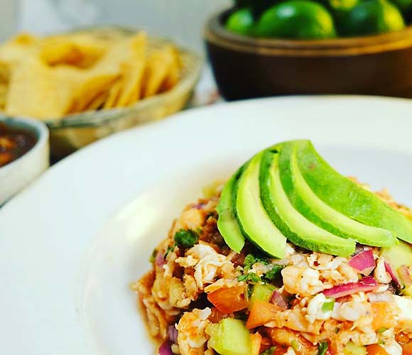 A rice dish is topped with avocado and is plated in front of tortilla chips and a basket of limes.