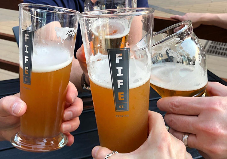 A cold, light beer in a pint glass labeled “Fife Street Brewing” sits on a wooden bar next to a house specialty sandwich partially unwrapped in foil.