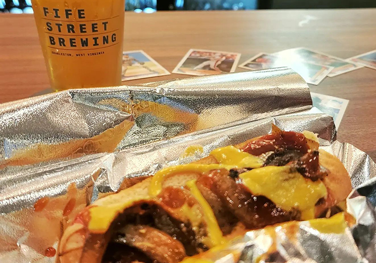 A cold, light beer in a pint glass labeled “Fife Street Brewing” sits on a black wooden table. A salmon-colored cocktail sits in the blurry background.