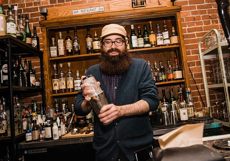 A bearded bartender in a black cartigan and cap, shakes a cocktail in a mixing glass. 