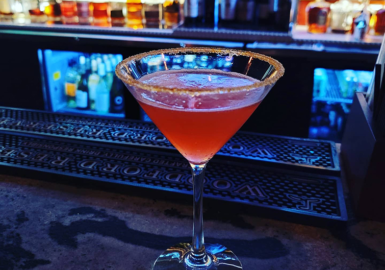 A raspberry-colored cocktail fills a sugar-rimmed martini glass that sits on a lounge bar. 