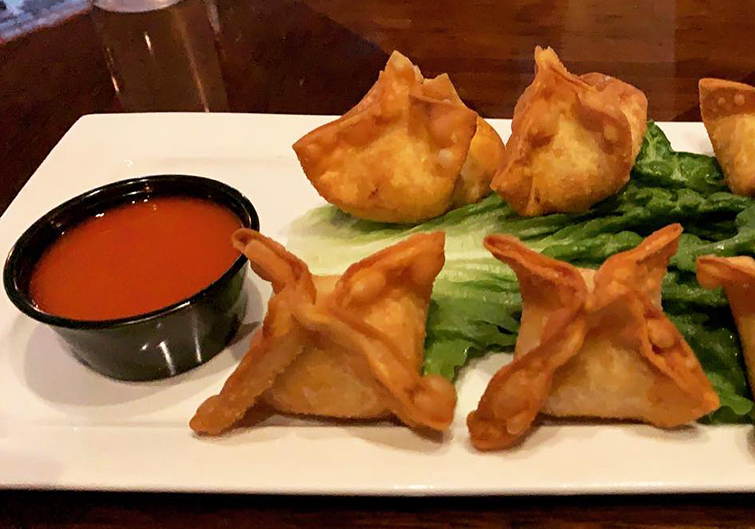 Six fried wontons sit atop lettuce leaves on a white plate with a red dipping sauce next to it. 