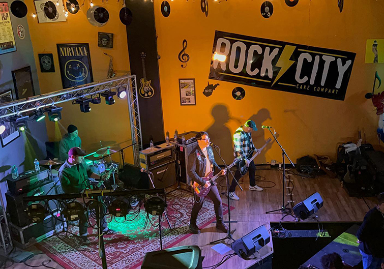 A rock band of four males are playing music inside the Rock City Cake Company in Charleston, West Virginia.