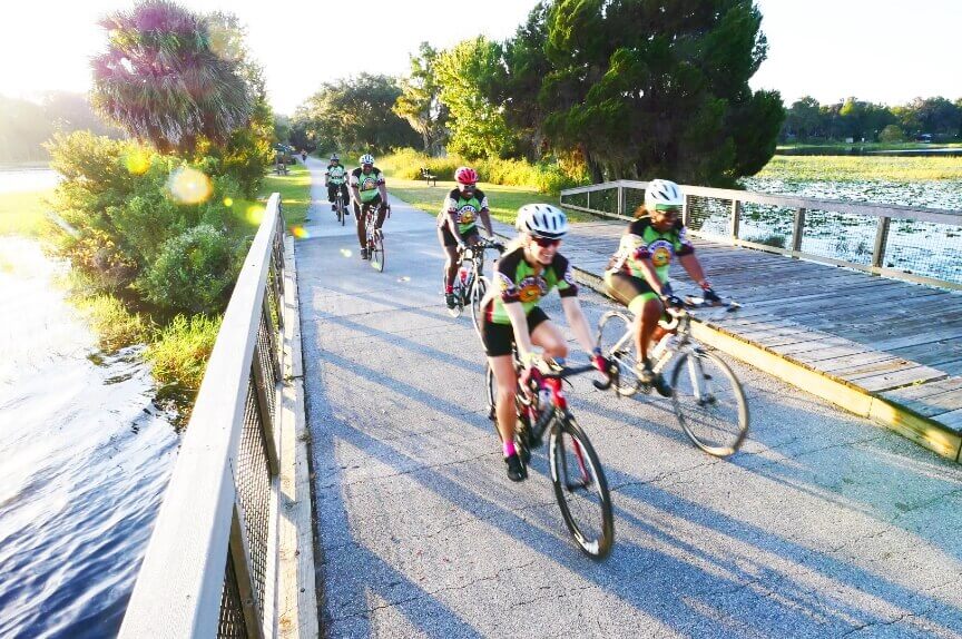 Large group of cyclists traversing a bridge over water while the sun shines behind them