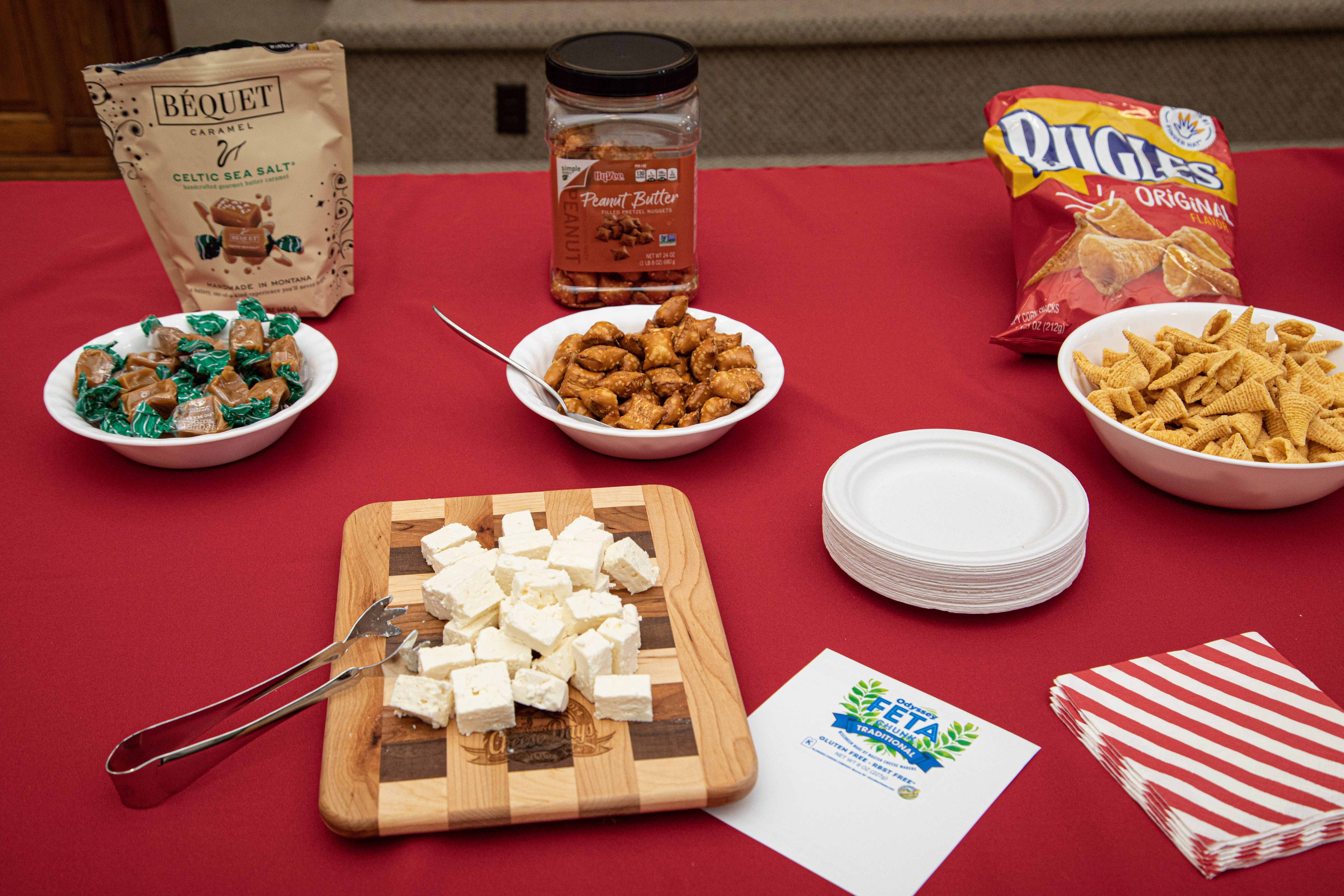 A red table holds a wooden chopping board with feta cheese chunks, a bowl of caramels, a bowl of peanut butter bites, a bowl of bugles snacks, a stack of plates and a stack of red and white striped napkins.