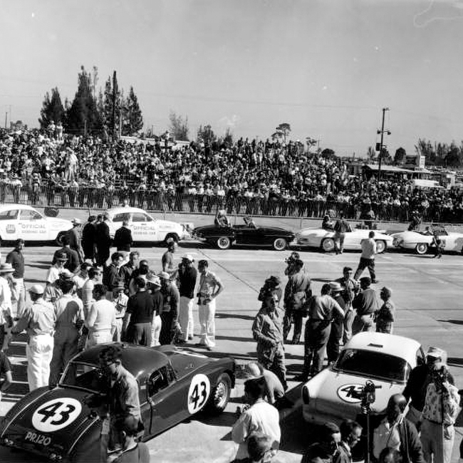 A black and white vintage photo of sportscars parked on the Sebring International Raceway while onlookers watch from the stands in Sebring, Florida.