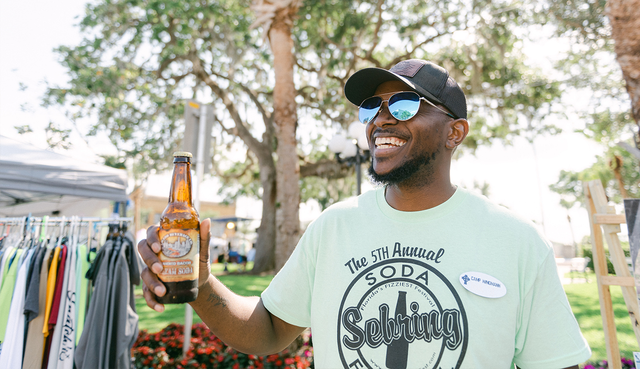 A Black male wearing a hat, glasses and a green shirt holds a brown bottle of craft soda at the Sebring Soda Festival in Sebring, Florida.
