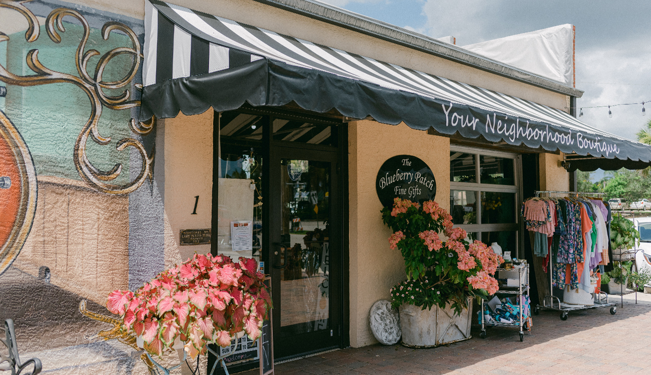 The charming storefront of The Blueberry Patch in Lake Placid, Florida is decorated with a striped black and white awning, pink flowers and racks of merchandise. 