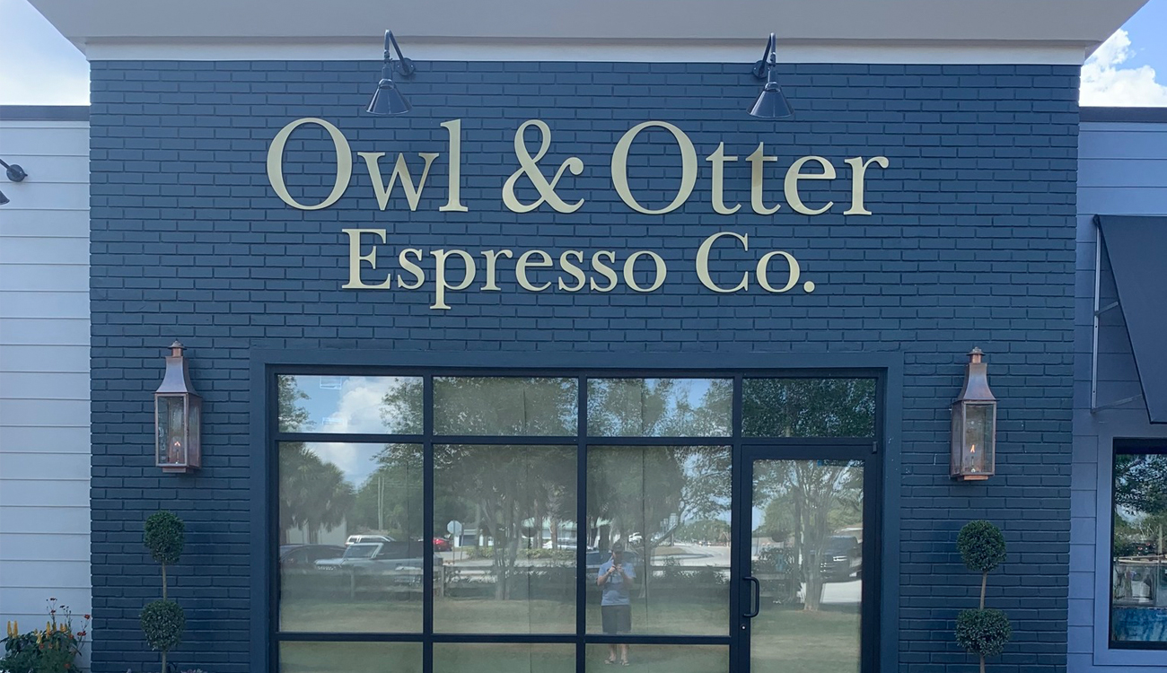 The outside storefront of Owl & Otter Espresso Co. in Lake Placid, Florida is painted two shades of blue with adorning lights on each side of glass windows.