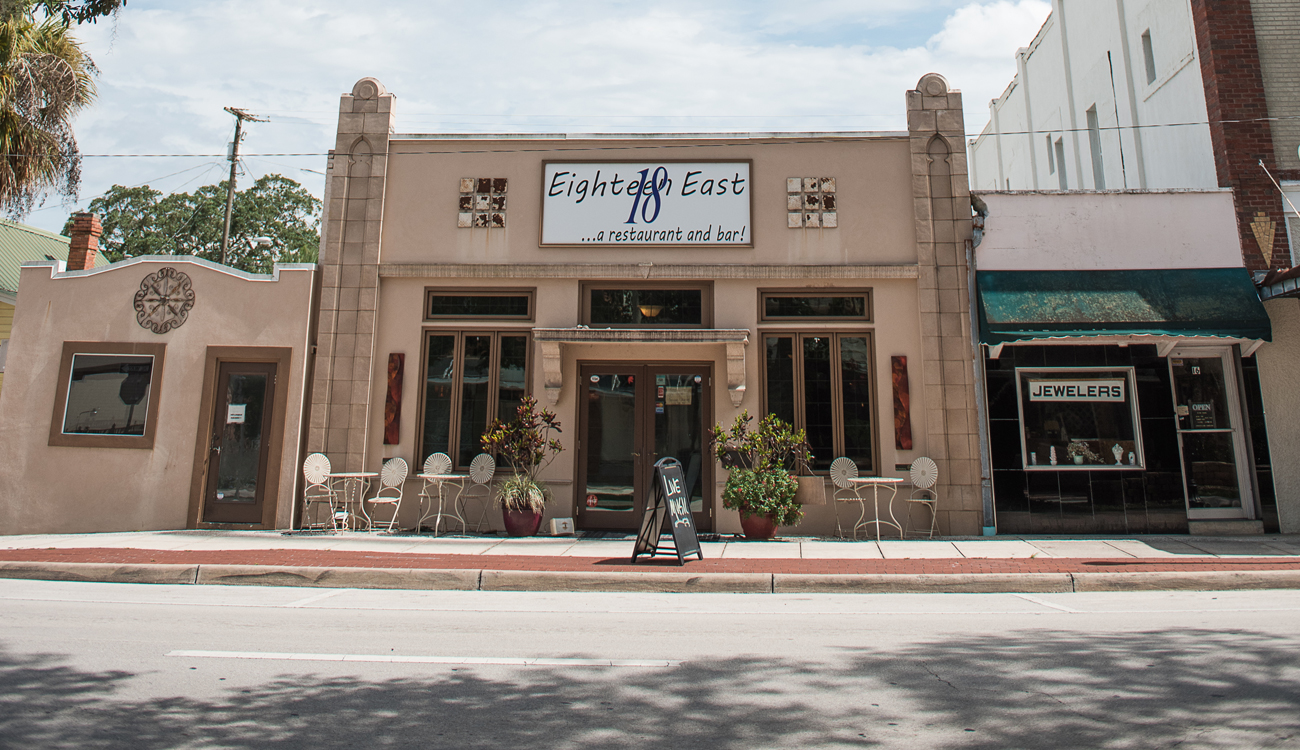 The quaint storefront of Eighteen East Restaurant & Bar in Avon Park, Florida is paiinted beige with cream outdoor patio residing outside.  