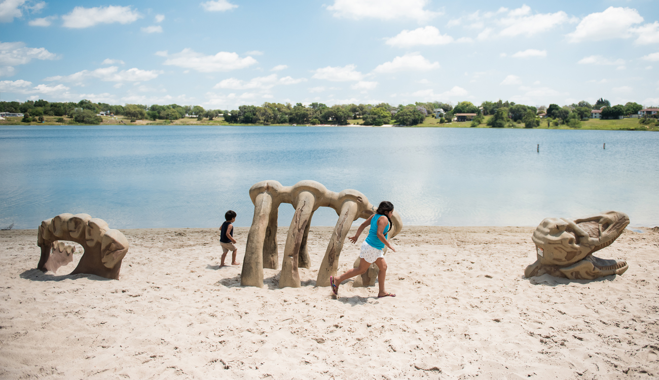 A sandy beachfront with play equipment shaped like a dinosaur skeleton is being enjoyed by two young children at Donaldson Park in Avon Park, Florida. 