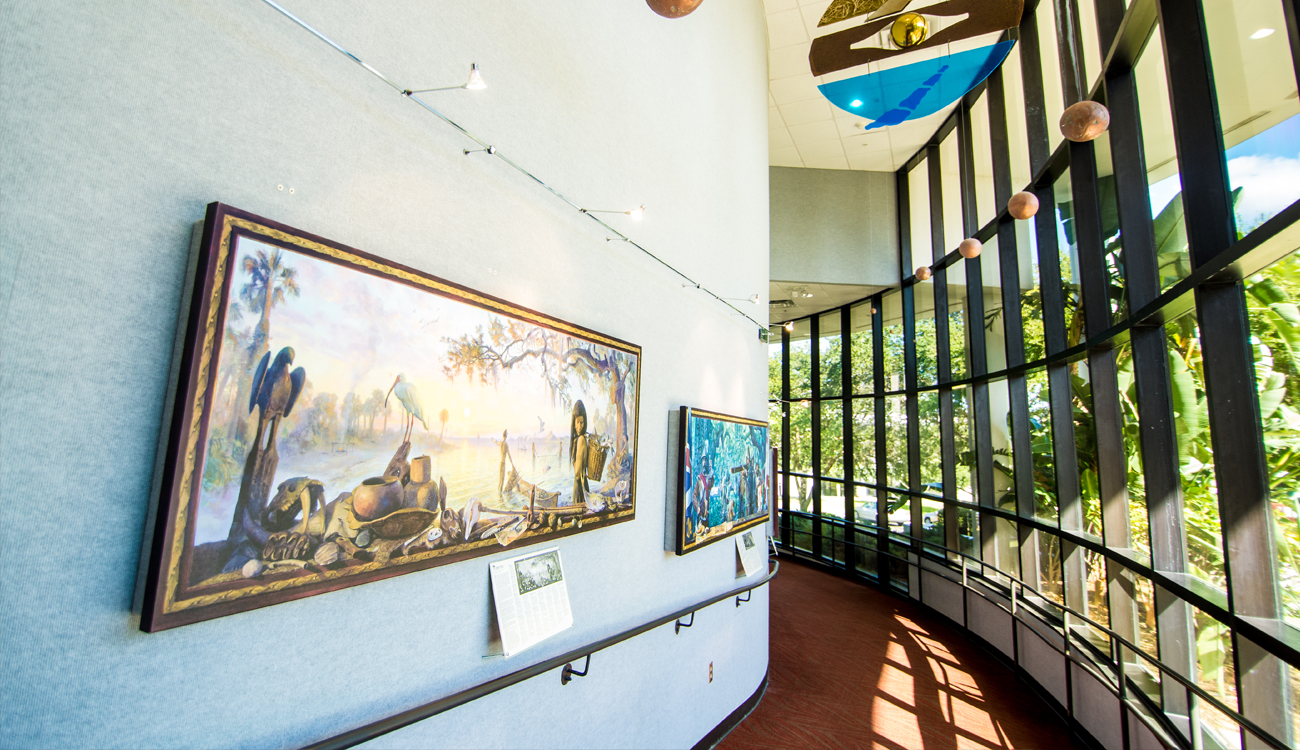 A curved walkway with glass windows on one side and hung artwork on the other side ushers guests to more exhibits in the Museum of Florida Art & Culture in Avon Park, Florida.