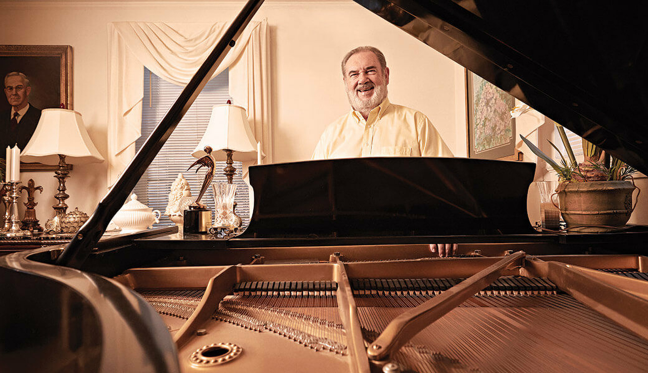 Composer Mark Andersen stands smiling behind a grand piano with the top lifted in an elegantly decorated room with paintings and window coverings in Lumberton, North Carolina.